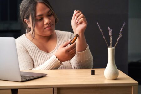 A woman uses a therapeutic essential oil roll-on at a work desk featuring a laptop and vase with flowers.